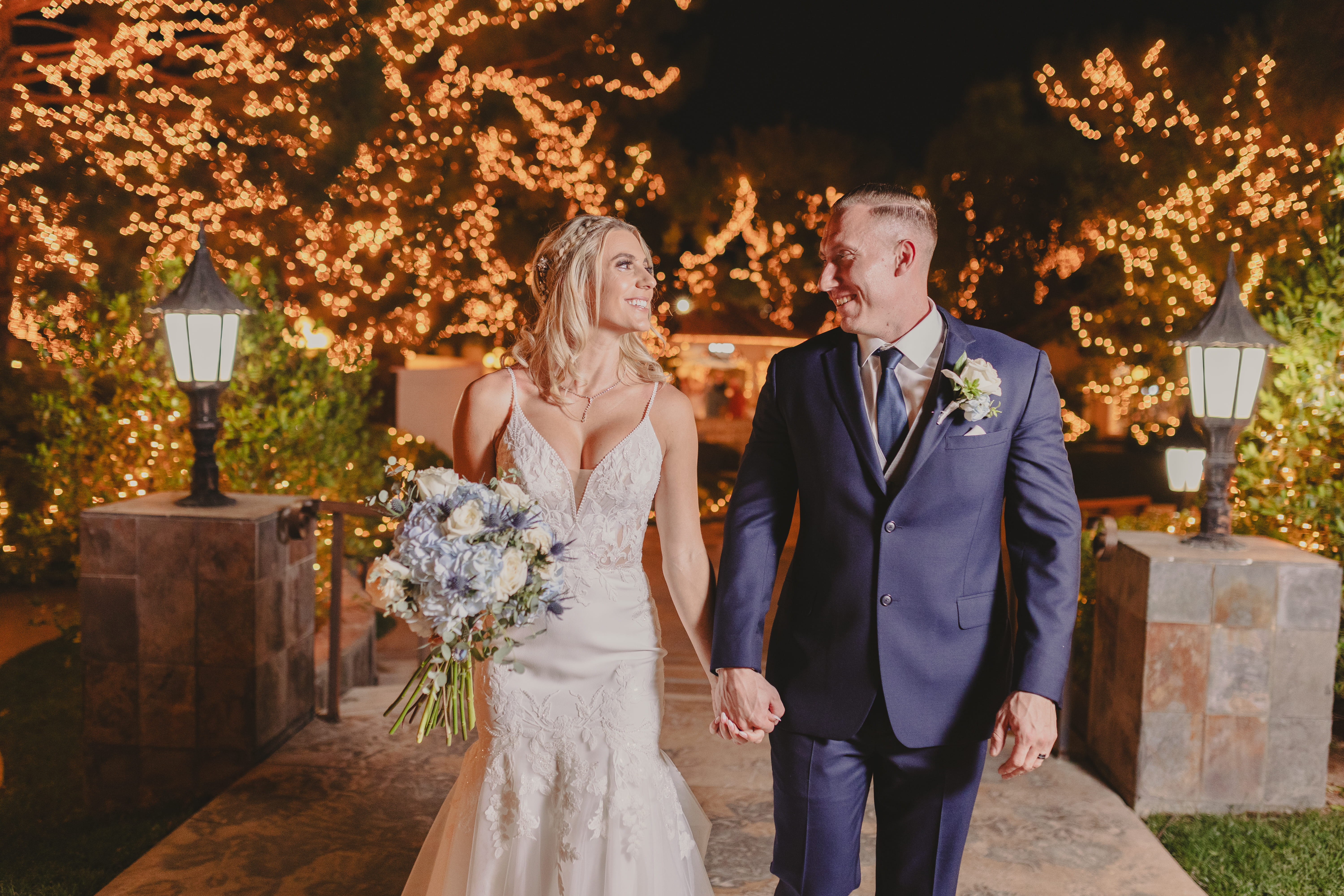 Newlyweds walking hand in hand outdoors at night under glowing string lights and illuminated trees in a Las Vegas wedding venue