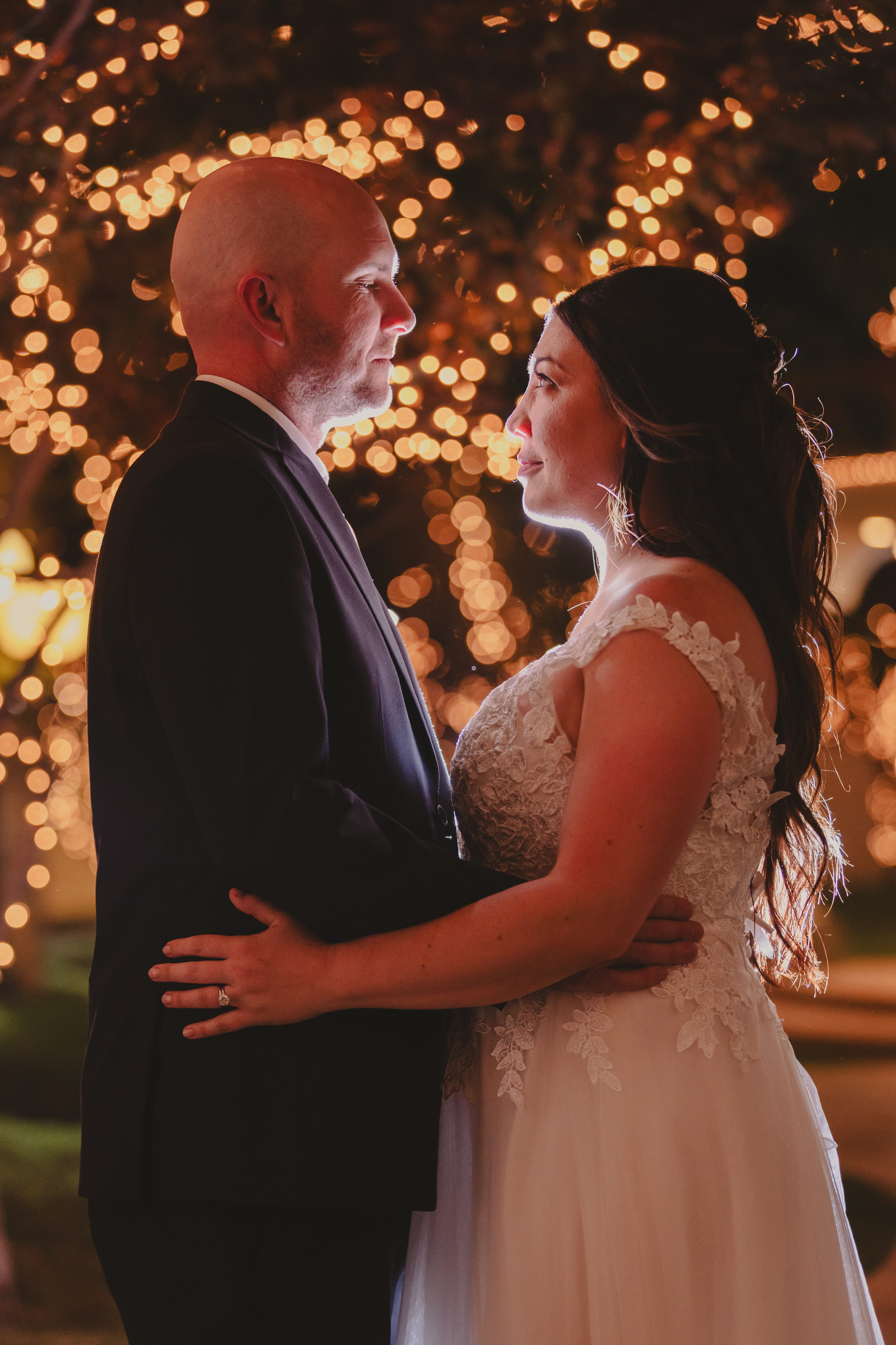 Bride and groom facing each other with warm string lights glowing behind them during a romantic evening wedding portrait