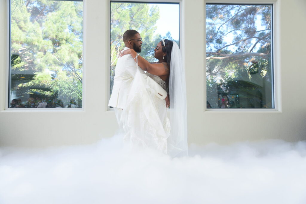 Bride and groom embracing in front of large sunlit windows inside a bright chapel, with soft fog effect creating a romantic wedding portrait
