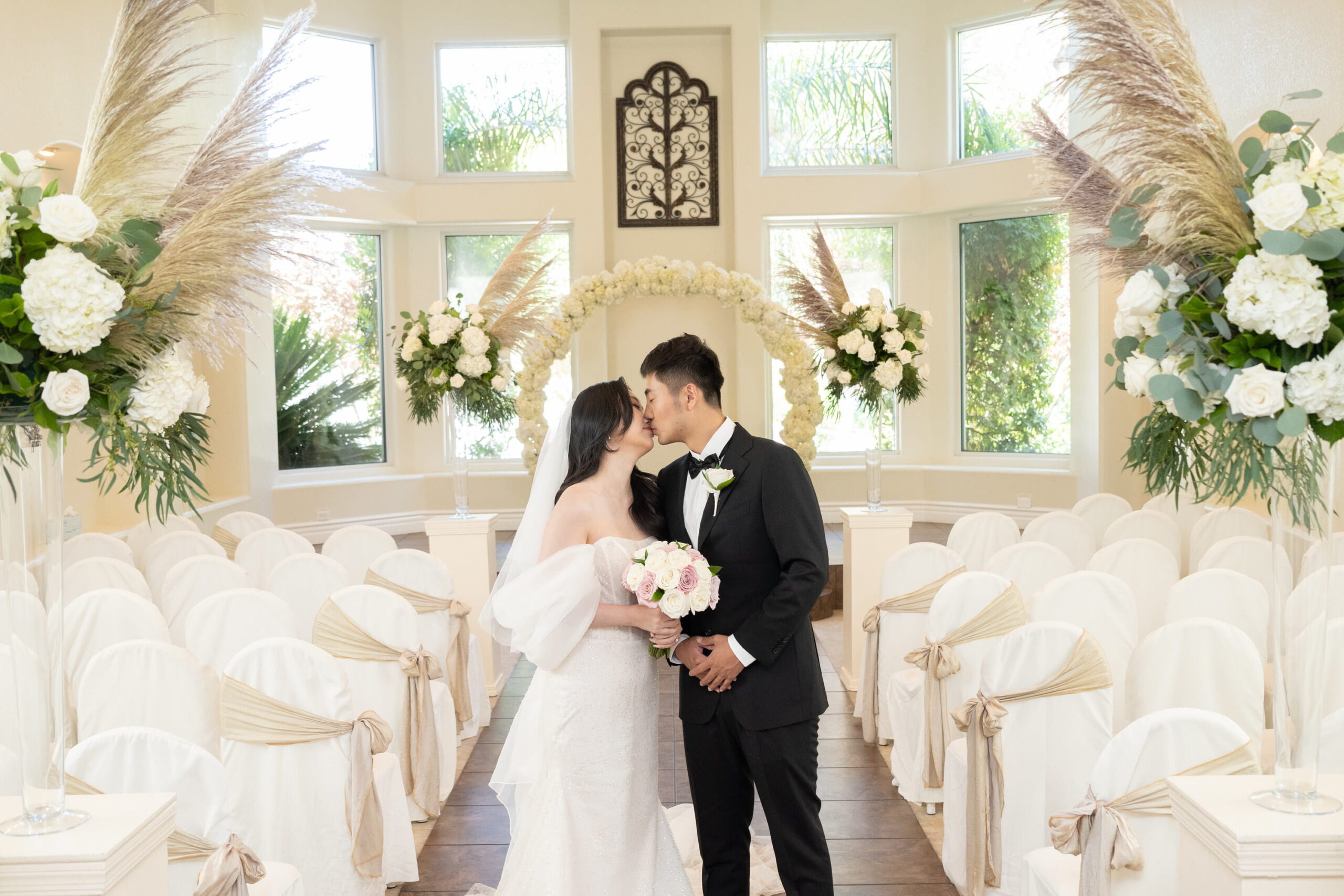 Bride and groom standing inside Sunset Gardens wedding chapel with floral arch, white seating, and natural light