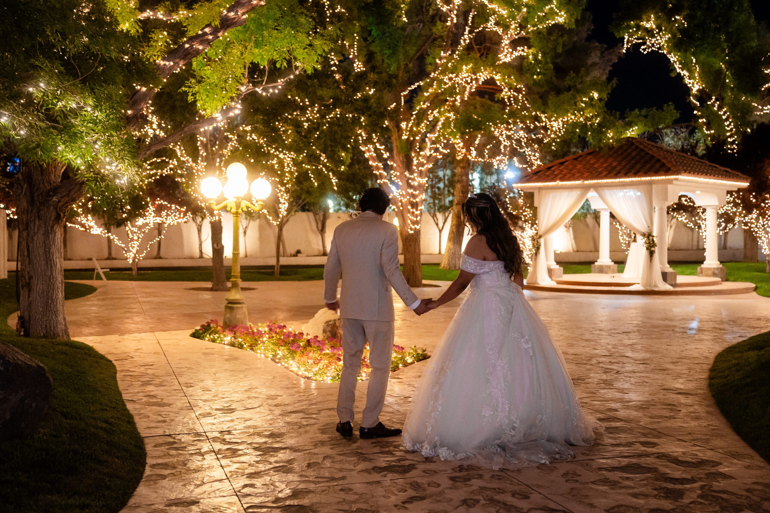 Bride and groom walking hand in hand under glowing string lights toward a gazebo at a romantic evening wedding venue