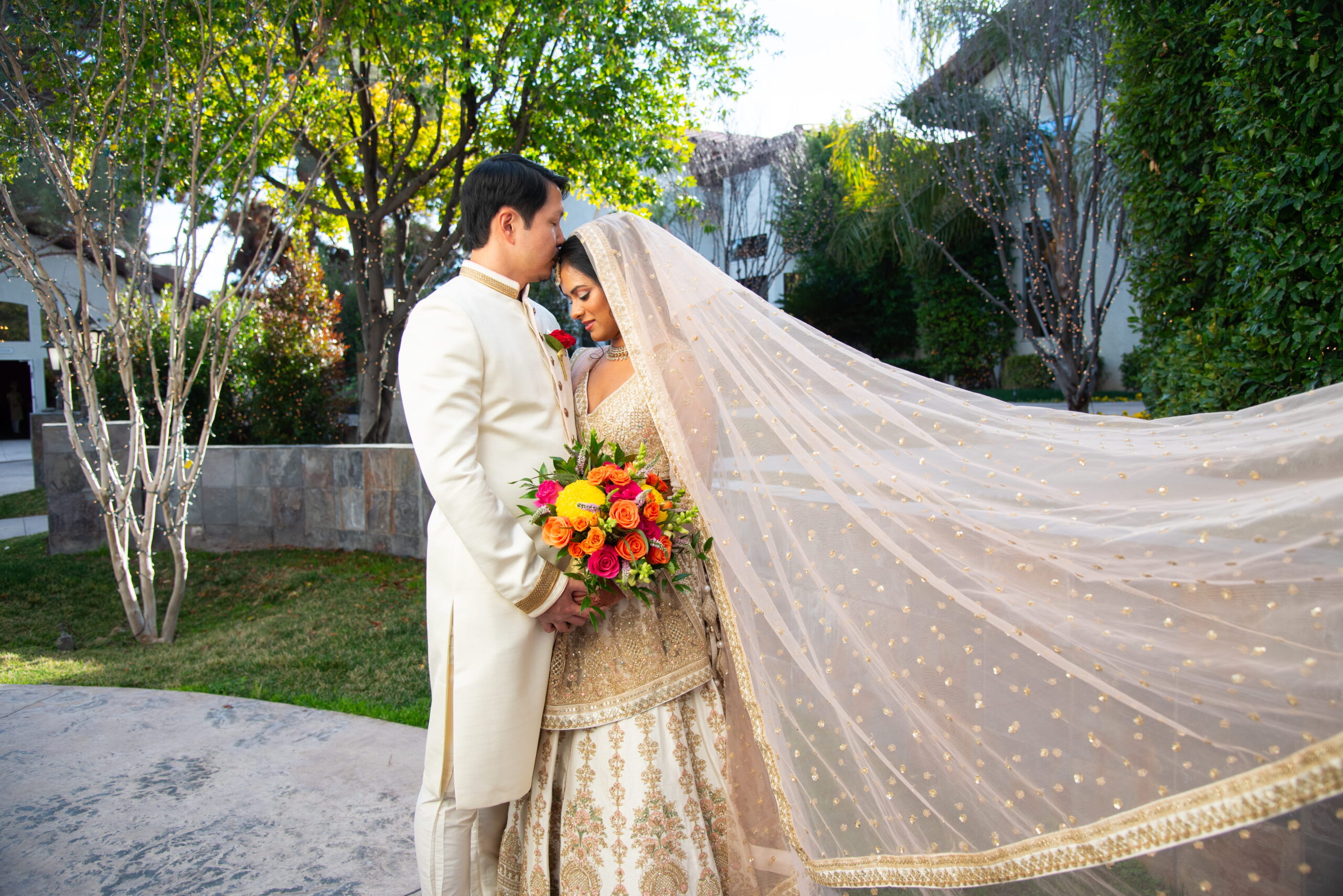 Bride and groom in traditional attire with a long flowing veil and vibrant bouquet during an outdoor wedding portrait
