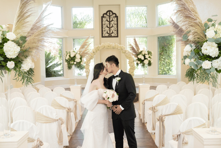 Bride and groom standing inside Sunset Gardens wedding chapel with floral arch, white seating, and natural light