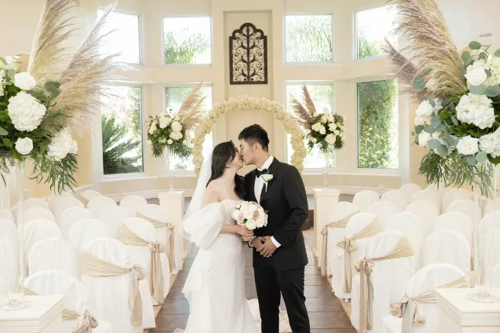 Bride and groom standing inside Sunset Gardens wedding chapel with floral arch, white seating, and natural light